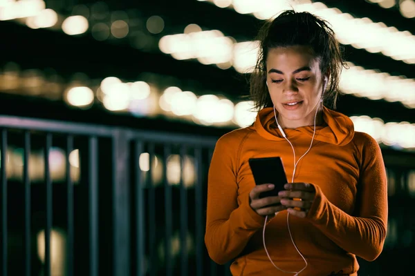 Geceleri telefonla dolaşan kulaklıklı genç bir sporcu kadın. Gece sporu, gece koşucusu, gece sporu.