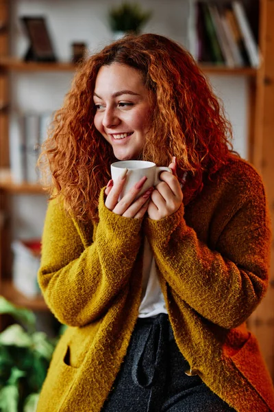 Kızıl saçlı bir kız sabah kahvesini evde içiyor. Sıcak evinde elinde sıcak bir fincan sabah kahvesi tutan mutlu bir kızıl kız..