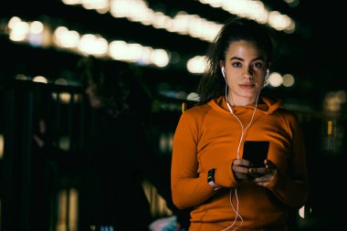 Kadın gece koşucusu karanlıkta duruyor ve dışarıda müzik dinlemek için telefon kullanıyor..
