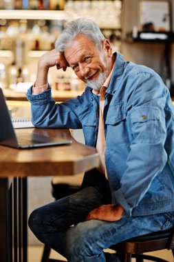 Modern son sınıf öğrencisi kafeteryada oturuyor, dizüstü bilgisayarla çalışıyor ve kameraya gülümsüyor..