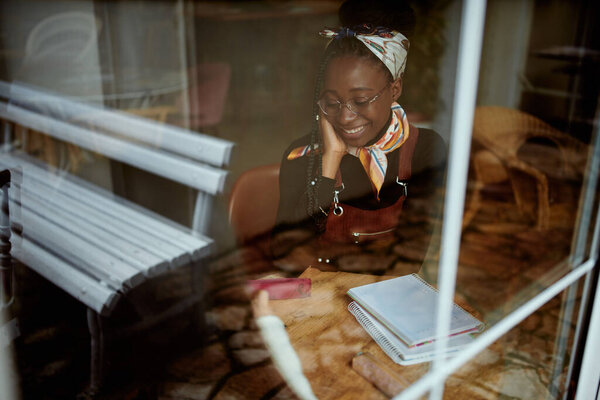 A smart African college girl sits in the cafeteria and studies for an exam. The picture was taken from the outside. A girl on student exchange sitting in a coffee shop and studying.