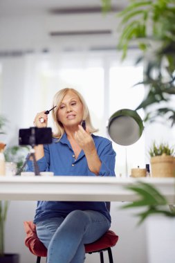 Gülümseyen son sınıf öğrencisi bir kadın güzellik köşesinde oturuyor ve maskara sürüyor..