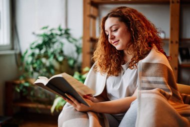 Oturma odasında battaniyeye sarılmış güzel bir kızıl kız sabahları ilginç bir kitap okuyor..