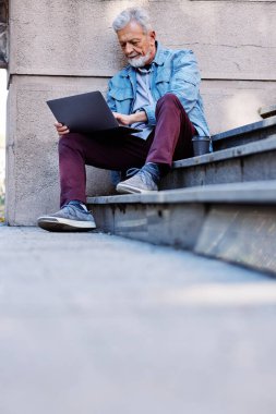 Üniversite binasının önündeki merdivenlerde elinde dizüstü bilgisayarla oturan son sınıf öğrencisi ders çalışıyor. Geri çekilen konsept.