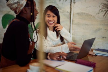 İki çok kültürlü üniversiteli kız kafeteryada duruyor ve araştırma için dizüstü bilgisayar kullanıyorlar. E-öğrenim ve veri erişilebilirliği.