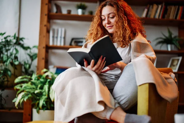 Oturma odasında battaniyeye sarılmış güzel bir kızıl kız sabahları ilginç bir kitap okuyor..
