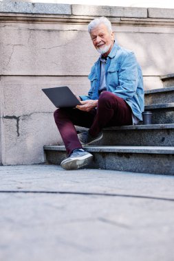 Üniversite binasının önündeki merdivenlerde elinde dizüstü bilgisayarla oturan mutlu bir son sınıf öğrencisi ders çalışıyor. Geri çekilen konsept.