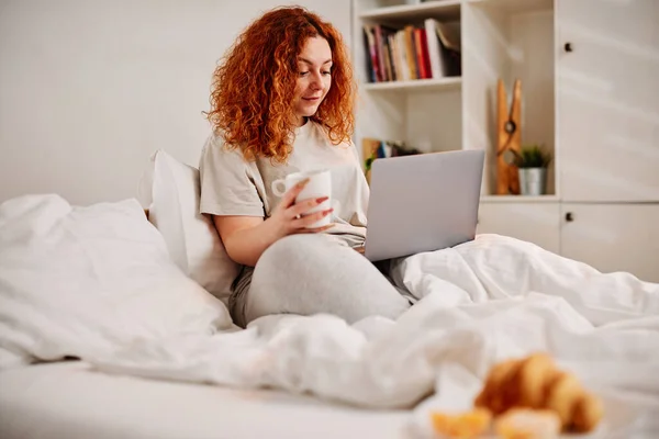 A ginger girl sits on the bed in the morning, drinking her morning coffee and using her laptop.