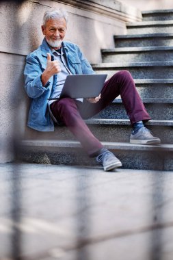 Mutlu bir son sınıf öğrencisi tuğla duvara yaslanmış dizüstü bilgisayarını tutuyor ve baş parmağını kaldırıyor..