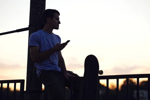 Genç Bir Çocuğun Silueti Kaykaylı Bir Korkulukta Oturuyor Telefonunu Kullanıyor — Stok fotoğraf