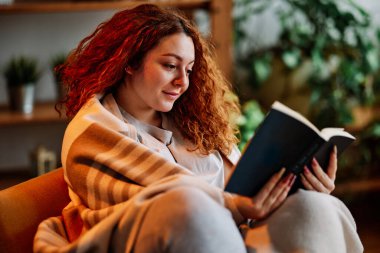 Oturma odasında battaniyeye sarılmış güzel bir kızıl kız sabahları ilginç bir kitap okuyor..