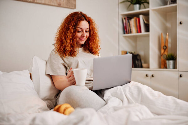 Рыжая девушка сидит на кровати по утрам, пьет утренний кофе и пользуется ноутбуком..