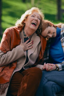 Bir büyükanne parkta bankta otururken kızıl ergen torunuyla gülüyor ve el ele tutuşuyor..