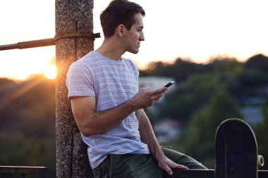 Genç bir çocuk kaykaylı bir korkulukta oturuyor ve telefonunu kullanıyor..