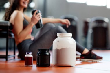 Spor salonunda arka planda sportif bir kadın olan protein tozu ve multivitamin kavanozlarına odaklanın..