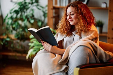 Kızıl kıvırcık saçlı genç bir kadın oturma odasında bir sandalyede otururken kitap okuyor..