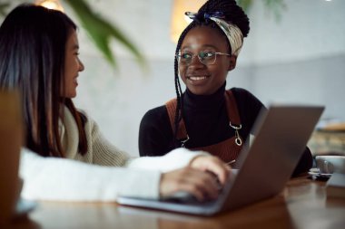 İki çok kültürlü kız öğrenci, kafeteryada kahve içerken dizüstü bilgisayarlarında proje yapıyor..