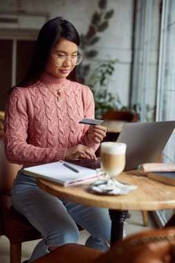 Genç, sevimli Asyalı kadın, kahve dükkanında cappuccino içerken bilgisayarında alışveriş yapıyor..