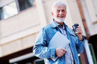 Yeniden eğitim programındaki mutlu bir son sınıf öğrencisi elinde bir laptop ve kahveyle üniversitenin önünde duruyor..