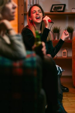 Genç bir kadın bir mikrofon tutuyor ve oturma odasında arkadaşıyla karaoke partisinde şarkı söylüyor..