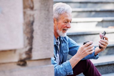 Üst sınıftan bir adam dışarıda merdivenlerde oturup kahve içiyor ve arkadaşları ve ailesiyle telefonda video görüşmesi yapıyor..