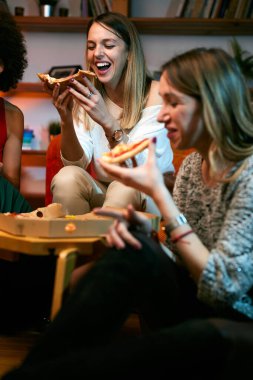 Çok kültürlü bir grup neşeli genç kadın oturma odasında otururken pizza yiyor..