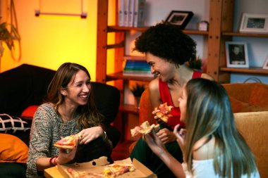 Çok kültürlü bir grup neşeli genç kadın oturma odasında otururken pizza yiyor..
