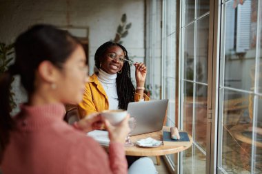 İki çok kültürlü kız öğrenci, kafeteryada kahve içerken dizüstü bilgisayarlarında proje yapıyor..