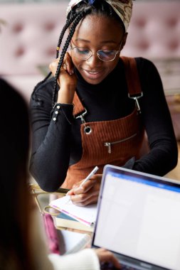 İki çok kültürlü kız öğrenci, kafeteryada kahve içerken dizüstü bilgisayarlarında proje yapıyor..