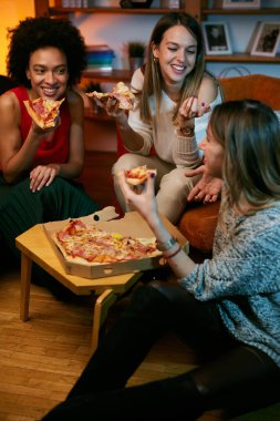 Çok kültürlü bir grup neşeli genç kadın oturma odasında otururken pizza yiyor..