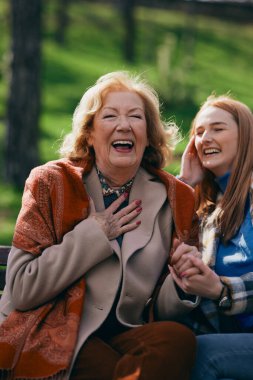 Bir büyükanne parkta bankta otururken ergen torunuyla gülüp eğleniyor..