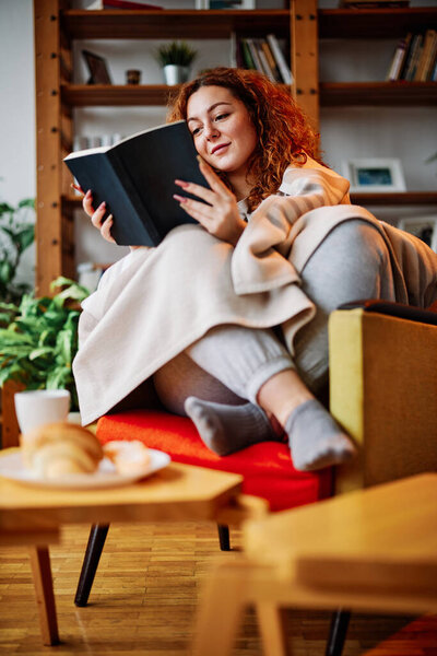 A young woman with red curly hair covered in the blanket is reading a book while sitting in a chair in her living room.