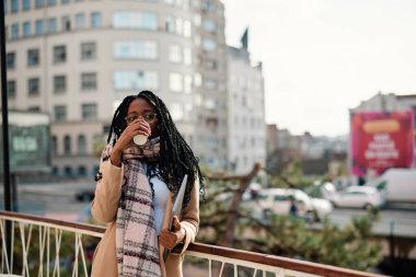 Neşeli, genç bir Afrikalı kadın kahvesini içerken bir kafenin önünde çitlere yaslanıyor..