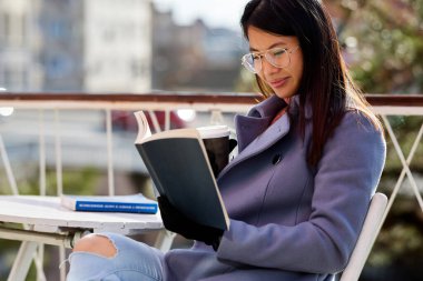 Genç Asyalı kadın kafenin dışında kahve içerken kitap okuyor..