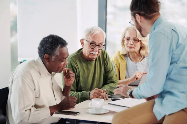 Genç bir eğitimci sınıftaki bir masada oturuyor ve son sınıf öğrencilerine web tasarımı anlatıyor. Ortadaki yaşlı adama odaklan..