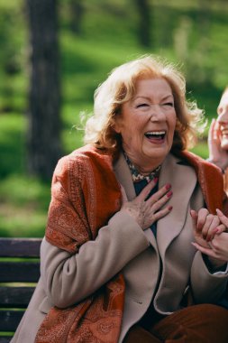 Genç bir kızıl kadın, soğuk havada büyükannesiyle oturuyor ve bankta konuşuyor..