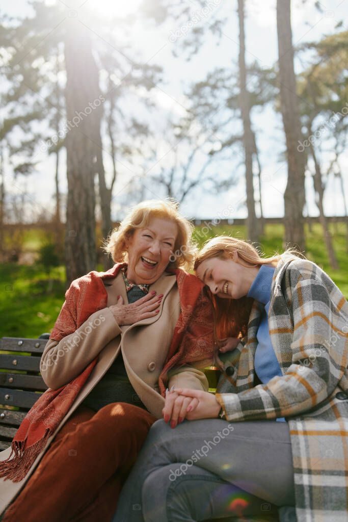 A grandmother is laughing and having fun with her adolescent granddaughter while sitting on the park bench.