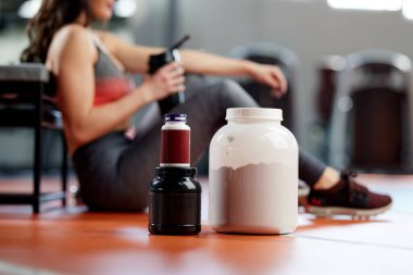 Spor salonunda arka planda sportif bir kadın olan protein tozu ve multivitamin kavanozlarına odaklanın..