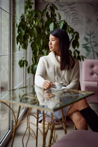A thoughtful asian woman is sitting in a coffee shop and looking trough the window while drinking a coffee.