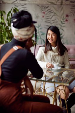 İki genç çok kültürlü kadın bir kafede pencerenin kenarında oturmuş kahve içiyorlar..