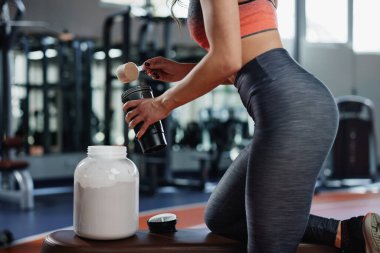 Kaslı bir sporcu kadın spor salonundaki bir bankta eğilir ve bardağına protein tozu karıştırır..