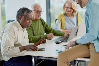 Genç bir profesör sınıftaki bir masada oturuyor ve çok kültürlü son sınıf öğrencilerine ders veriyor..