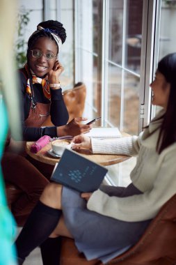 İki genç çok kültürlü kadın bir kafede pencerenin kenarında oturmuş kahve içiyorlar..