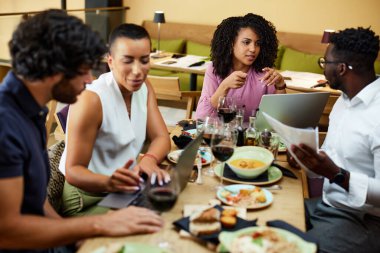 A group of multicultural managers is sitting in a restaurant at a dinner table, talking about a project and using technologies to improve it.