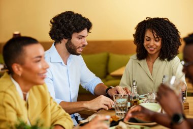 A multicultural group of friends sit in a restaurant and eat dinner.
