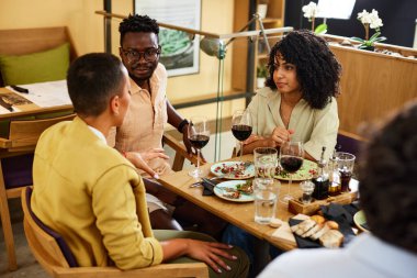 A group of multicultural friends sits in a restaurant and chat after dinner.
