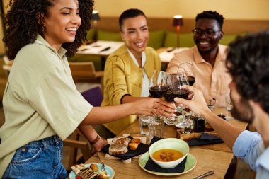 A small group of multicultural friends is sitting in a restaurant and toasting with red wine.