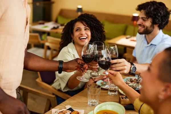 A small group of multicultural friends is sitting in a restaurant and toasting with red wine.