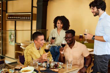 A happy multiracial group of friends celebrates the event in a restaurant. They are drinking wine and laughing.