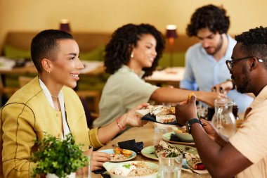 A cheerful woman is holding a plate with food and sharing it with her friend in a restaurant at the dinner table.